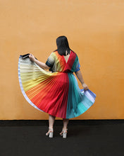 Cargar imagen en el visor de la galería, A woman viewed from behind wearing a pleated rainbow-striped dress and white high heels, holding out both sides of the skirt to show the full color spectrum against a solid orange wall.