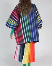 Cargar imagen en el visor de la galería, A model shown from the back wearing a vibrant, oversized striped quilted coat and a matching pleated skirt with rainbow colors, set against a plain light gray studio background.