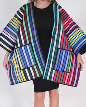 Cargar imagen en el visor de la galería, A model wearing a vibrant, quilted jacket with bold vertical stripes in multiple colors including blue, red, yellow, green, pink, and white, styled over a black dress against a light gray background.